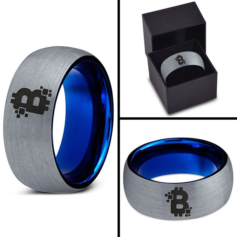Op de afbeelding: Een zilveren ring met een blauwe binnenband en een zwart Bitcoin-symbool gegraveerd op de bovenkant. De ring zit in een zwarte doos.