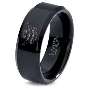 May include: A black tungsten carbide ring with a beveled edge. The ring features a detailed engraving of a barrel with wings. The words "Tungsten Carbide" are etched on the inside of the ring.