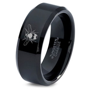 May include: A black tungsten carbide ring with a beveled edge. The ring features a detailed engraving of a bee on the front. The words "TUNGSTEN CARBIDE" are printed on the inside of the ring.