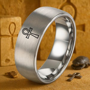 May include: A silver-colored ring with a brushed metal finish. The ring features a black ankh symbol on the outer band. The ring is displayed on a textured, tan surface with other Egyptian-themed objects.