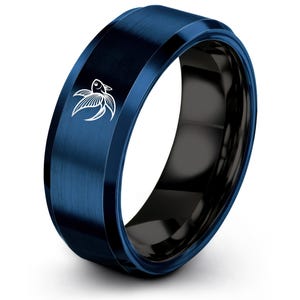 Puede incluir: Un anillo azul con interior negro. El anillo tiene un acabado de metal cepillado y un diseño de pez blanco en el lateral. El anillo es de estilo moderno y probablemente está hecho de metal.