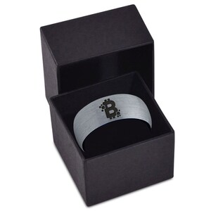 Op de afbeelding: Een zilveren ring met een zwart Bitcoin-symbool dat erop is gegraveerd. De ring zit in een zwarte fluwelen doos.