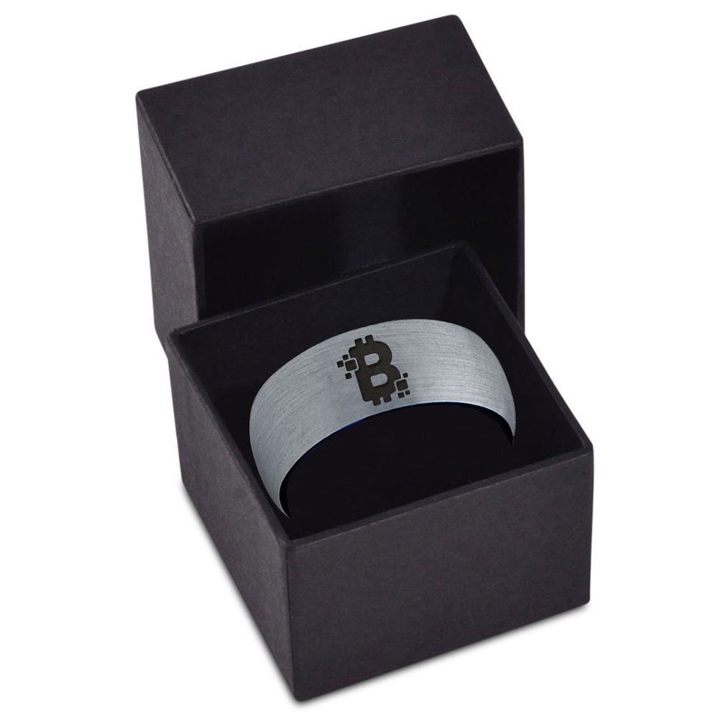 Op de afbeelding: Een zilveren ring met een zwart Bitcoin-symbool dat erop is gegraveerd. De ring zit in een zwarte fluwelen doos.