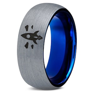 May include: A silver tungsten carbide ring with a blue inner band and a rocket ship design engraved on the outside.