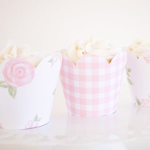 May include: Three cupcakes with white frosting and pink and white patterned cupcake liners. The liners feature a gingham pattern and a floral pattern with pink roses.