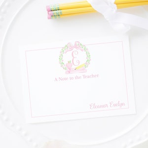 Può includere: Un biglietto bianco con una ghirlanda floreale rosa e la lettera "E" al centro. La ghirlanda include una mela rosa e una matita. Il testo "A Note to the Teacher" è sotto la ghirlanda. Il nome "Eleanor Evelyn" è in fondo al biglietto.