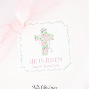 Op de afbeelding: Een wit cadeau-etiket met een groene bloemrand en een roze bloemkruis in het midden. De tekst "He is Risen" is in zwart onder het kruis gedrukt, gevolgd door "Love, the Thomas Family".