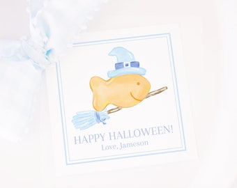 以下が含まれることがあります： 青いオレンジのハロウィーンギフトタグ。魔女の帽子をかぶってほうきに乗った、漫画の金魚が描かれています。タグには「ハッピーハロウィン！愛を込めて、ジェイムソン」と書いてあります。