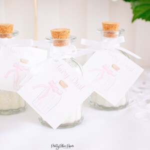 May include: Three small glass jars with cork tops filled with white glitter and tied with white ribbon. Each jar has a white tag with pink lettering that reads "Fairy Dust".