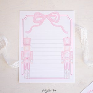K&ouml;nnte beinhalten: Rosa und wei&szlig;es bedruckbares Briefpapier mit zwei Nussknacker-Soldaten und einer rosa Schleife oben. Das Papier hat liniertes Schreibfeld und den Text "Pretty Plain Paper" unten.