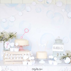 May include: A white table with a white tablecloth and a white cake on a pink cake stand. There are white cupcakes with bubble wands on top of them. There is a glass jar with a sign that says "Bubbles". The background is a white wall with blue and white watercolor bubbles.