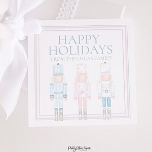 May include: A white gift tag with blue and pink borders. The tag says "Happy Holidays from the Lucas Family" and features three watercolor nutcracker illustrations.