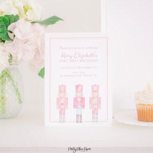 May include: A pink and white invitation card with three watercolor nutcracker soldiers. The card reads "Please join us as we celebrate Mary Elizabeth's VERY FIRST BIRTHDAY!" A cupcake with white frosting sits on a white plate.