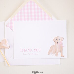K&ouml;nnte beinhalten: Eine wei&szlig;e Dankeskarte mit einem rosa karierten Umschlag. Die Karte zeigt eine Aquarell-Illustration eines Golden Retriever-Welpen mit einer rosa Schleife. Der Text "THANK YOU" und "Love, Sarah Anne" ist in rosa auf der Karte gedruckt.