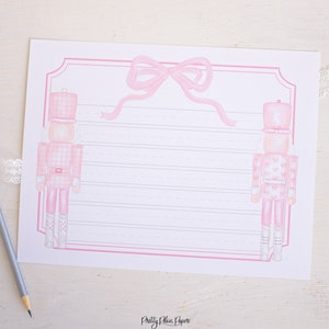 May include: Pink and white printable stationery featuring two pink nutcracker soldiers with a pink bow at the top. The paper has pink lines for writing.