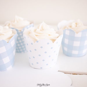 May include: Four cupcakes with white frosting in blue and white patterned cupcake liners. Two liners have a gingham pattern and two liners have a polka dot pattern.