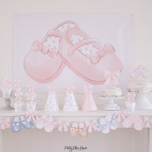 May include: A pink and white baby shower party table setting with a banner featuring a pair of pink baby shoes, cupcakes, cake pops, and a cake with a pink baby shoe topper. The table is decorated with a pink and white garland of baby shoes.