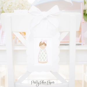 May include: White chair with a white bow and a white frame with a pink and green illustration of a girl wearing a dress. The frame has a scalloped edge and the text "Pretty Plain Paper" is written below the frame.