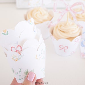 Puede incluir: Envoltorios blancos para cupcakes con borde festoneado, decorados con lazos de acuarela y detalles florales. Algunos envoltorios están vacíos, mientras que otros contienen cupcakes con glaseado en espiral. Se ve un adorno rosa de lazo para cupcakes.