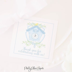May include: A blue and white cuckoo clock with a yellow bird on top. The clock is surrounded by green leaves and flowers. The text "Thank you for celebrating with us!" is printed below the clock. The text "Pretty Plain Paper" is printed at the bottom of the card.