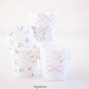 May include: Three cupcake liners with floral patterns. The top liner has a white background with a light green, yellow, and pink floral pattern. The middle liner has a white background with a light blue, yellow, and pink floral pattern. The bottom liner is white with a pink bow.