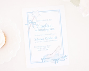 May include: A light blue invitation card with a blue border. The card features a watercolor illustration of a glass slipper, a magic wand, and a blue bow. The text on the card reads "Bibbidi Bobbidi Boo Caroline is turning two Please join us to celebrate Saturday, October 5th at 1 o'clock in the afternoon The Henderson Home 282 First Street Anderson, NC Pretty Plain Paper".