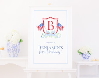 May include: A white sign with a blue and red shield with the letter "B" in the center. Two small American flags are on either side of the shield. The text "Welcome to Benjamin's first birthday!" is below the shield. The sign is on a white easel.