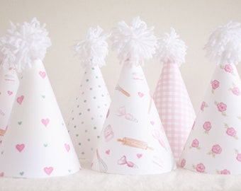 May include: Set of six pink and white party hats with different patterns, including hearts, polka dots, gingham, and floral designs. Each hat has a white pom-pom on top.