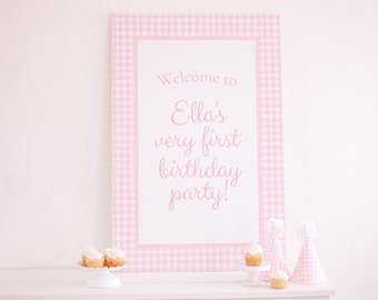 May include: Pink and white gingham welcome sign with the text "Welcome to Ella's very first birthday party!"  The sign is on a white surface with cupcakes and party hats.