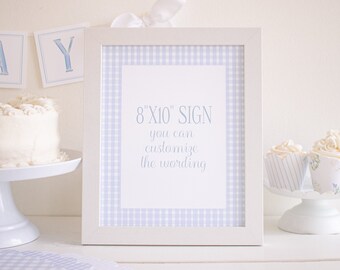 May include: A white frame with a blue and white gingham border surrounds a white sign with the text "8 x 10" SIGN you can customize the wording" in blue script. The sign is on a white surface with a white cake and cupcakes.