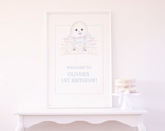 May include: White framed poster with a cartoon illustration of an egg with legs and arms sitting on a brick wall. The text on the poster reads "Welcome to Oliver's 1st Birthday!"