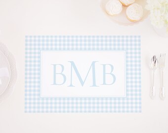 May include: A light blue gingham placemat with a white border and the monogram "BMB" in the center. The placemat is on a white table with a silver fork and spoon.