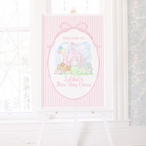 May include: A pink and white striped welcome sign with the text "Welcome to Lilibel's Three Ring Circus." The sign features a watercolor illustration of a circus tent, animals, and balloons, framed by a pink bow and oval border.