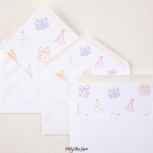 Pode incluir: Três envelopes brancos com um padrão de caixas de presente rosa e azul e chapéus de festa amarelos. Os envelopes estão aninhados um dentro do outro.