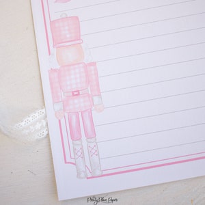 K&ouml;nnte beinhalten: Rosa-wei&szlig;er Gingham-Muster-Nussknacker mit wei&szlig;en Stiefeln und einer rosa Schleife. Der Nussknacker befindet sich auf rosa-wei&szlig;em, liniertem Papier mit dem Text "Pretty Plain Paper" unten.