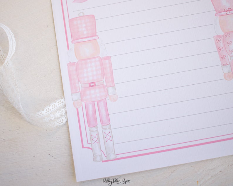 K&ouml;nnte beinhalten: Rosa-wei&szlig;er Gingham-Muster-Nussknacker mit wei&szlig;en Stiefeln und einer rosa Schleife. Der Nussknacker befindet sich auf rosa-wei&szlig;em, liniertem Papier mit dem Text "Pretty Plain Paper" unten.