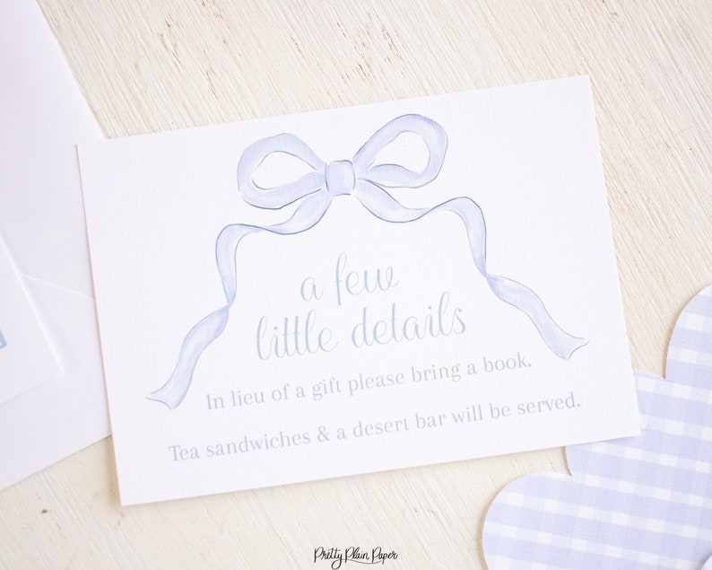May include: A white card with a blue ribbon bow and the text "a few little details" in cursive. The text below reads "In lieu of a gift please bring a book. Tea sandwiches & a desert bar will be served." The card is on a white wooden surface.