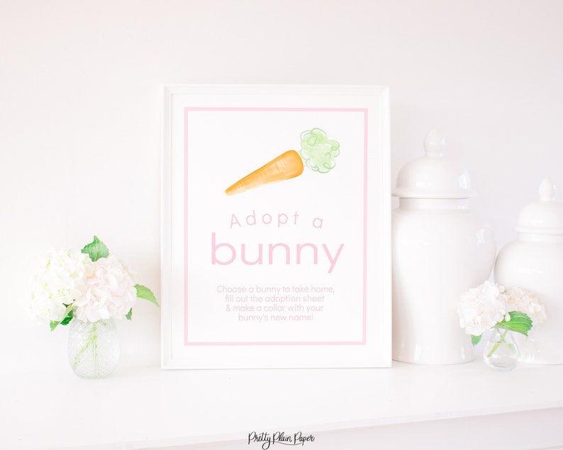 May include: A white framed sign with pink text that says "Adopt a bunny". A watercolour illustration of a carrot with green leafy tops is above the text. The sign is on a white shelf with white flowers in vases on either side.