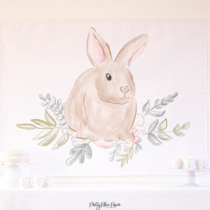 May include: A watercolor illustration of a brown bunny rabbit sitting in a wreath of green and blue leaves. The bunny is surrounded by pink flowers. The illustration is on a white background.