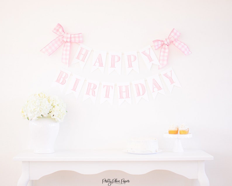May include: Pink and white "Happy Birthday" banner with gingham bows on a white background. A white table with a white cake and cupcakes is in the foreground.