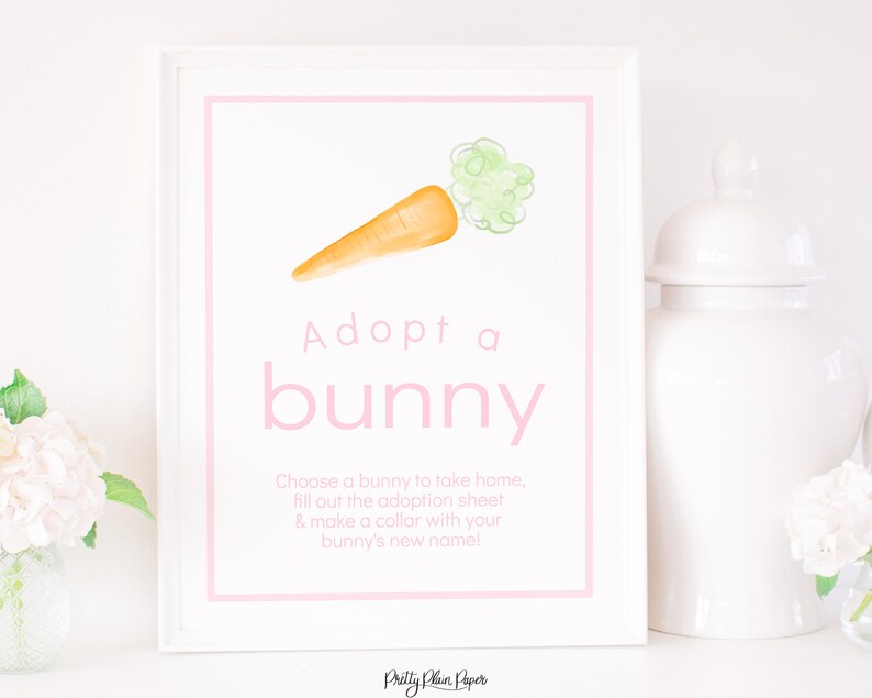 May include: A white sign with pink text that says "Adopt a bunny". There is a watercolour illustration of a carrot with green leaves on the sign. The text below the title says "Choose a bunny to take home, fill out the adoption sheet & make a collar with your bunny's new name!"