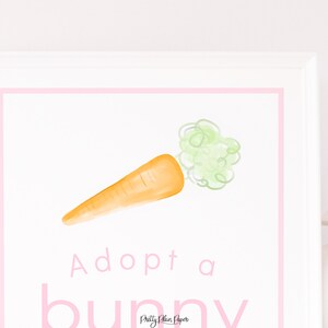 May include: A white framed print with a pink border. The print features a watercolour illustration of a carrot with green leafy tops. The text "Adopt a bunny" is written in pink below the carrot.