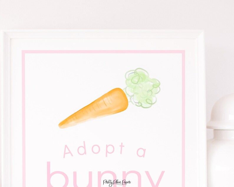 May include: A white framed print with a pink border. The print features a watercolour illustration of a carrot with green leafy tops. The text "Adopt a bunny" is written in pink below the carrot.