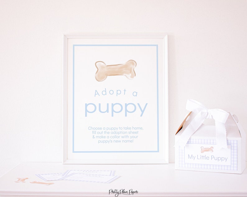 May include: A white framed sign with blue text that reads "Adopt a puppy". The sign features a brown bone illustration and instructions for a puppy adoption activity. A white gift box with a blue and white gingham pattern and a bone illustration is labeled "My Little Puppy".