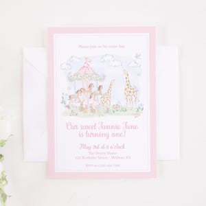 May include: A pink invitation card with a carousel and giraffe illustration. The text reads "Our sweet Tannie June is turning one!" and includes event details. The card is surrounded by a white envelope.