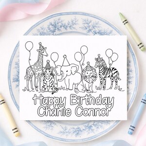 Op de afbeelding: Een witte verjaardagskaart met een lijntekening van een giraffe, tijger, olifant, leeuw en zebra, elk met een feesthoedje en ballonnen. De tekst "Happy Birthday Charlie Connor" staat eronder. De kaart ligt op een decoratief bord.