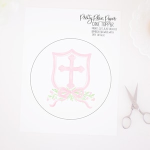 Könnte beinhalten: Druckbarer Tortendekor mit einem rosafarbenen Schild mit einem weißen Kreuz und einer rosafarbenen Schleife mit grünen Blättern. Der Text "Pretty Plain Paper CAKE TOPPER" befindet sich oben auf der Seite. Der Text "PRINT, CUT, & ATTACH TO BAMBOO SKEWER, WITH TAPE OR GLUE" befindet sich unterhalb des Tortendekors.