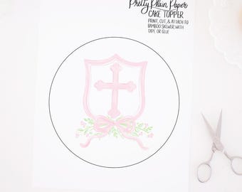 May include: Printable cake topper featuring a pink shield with a white cross and a pink bow with green leaves. The text "Pretty Plain Paper CAKE TOPPER" is at the top of the page. The text "PRINT, CUT, & ATTACH TO BAMBOO SKEWER, WITH TAPE OR GLUE" is below the cake topper.