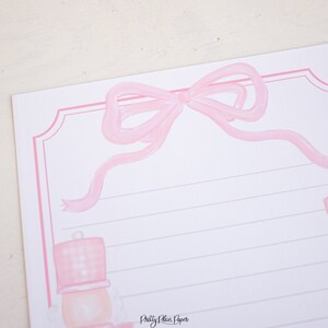 K&ouml;nnte beinhalten: Rosa Briefpapier mit einer rosa Schleife oben und einer rosa Nussknacker-Illustration unten. Das Papier hat liniertes Schreibfeld und den Text "Pretty Plain Paper" unten.