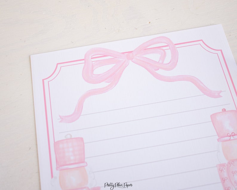 K&ouml;nnte beinhalten: Rosa Briefpapier mit einer rosa Schleife oben und einer rosa Nussknacker-Illustration unten. Das Papier hat liniertes Schreibfeld und den Text "Pretty Plain Paper" unten.